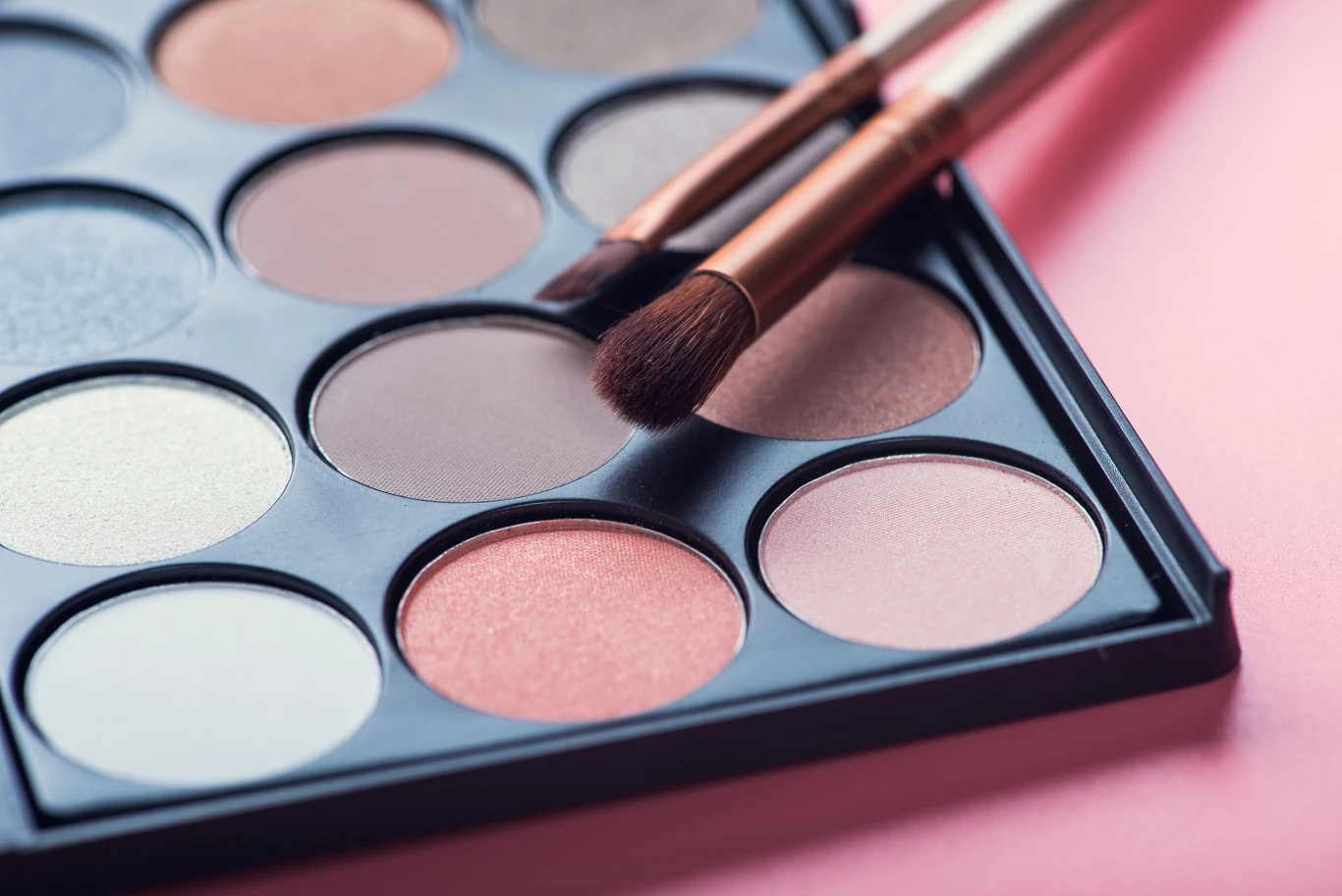 Paleta cieni do powiek w różnych odcieniach brązu, różu i beżu z dwoma pędzlami do makijażu ułożonymi na górze palety, całość na jasnoróżowym tle.