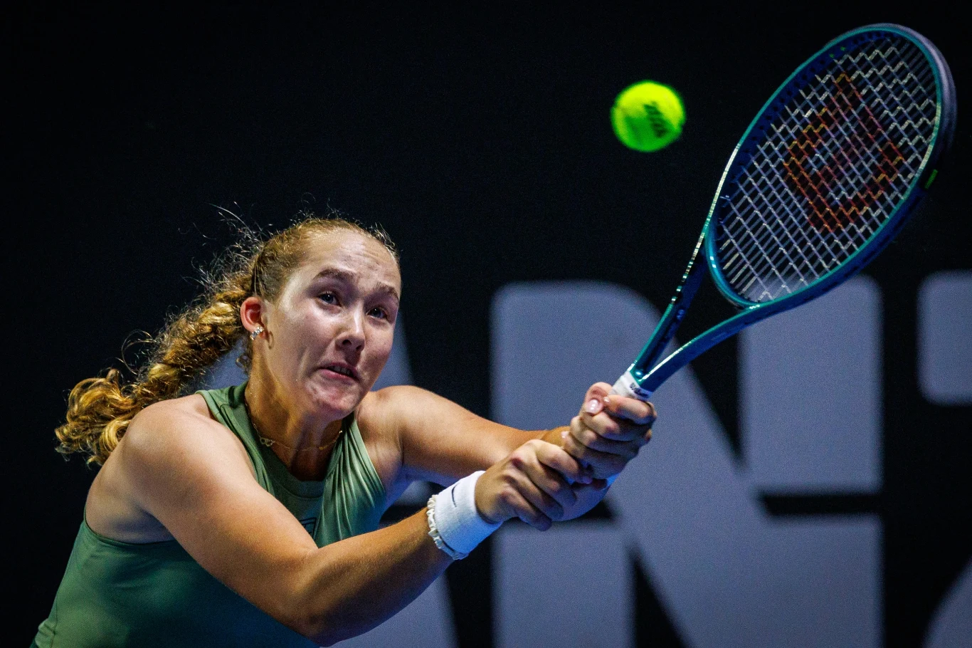 Tenisistka wykonuje uderzenie oburącz podczas dynamicznego meczu tenisowego, koncentrując się na nadlatującej piłce, światło podkreśla jej zaangażowanie i wysiłek.