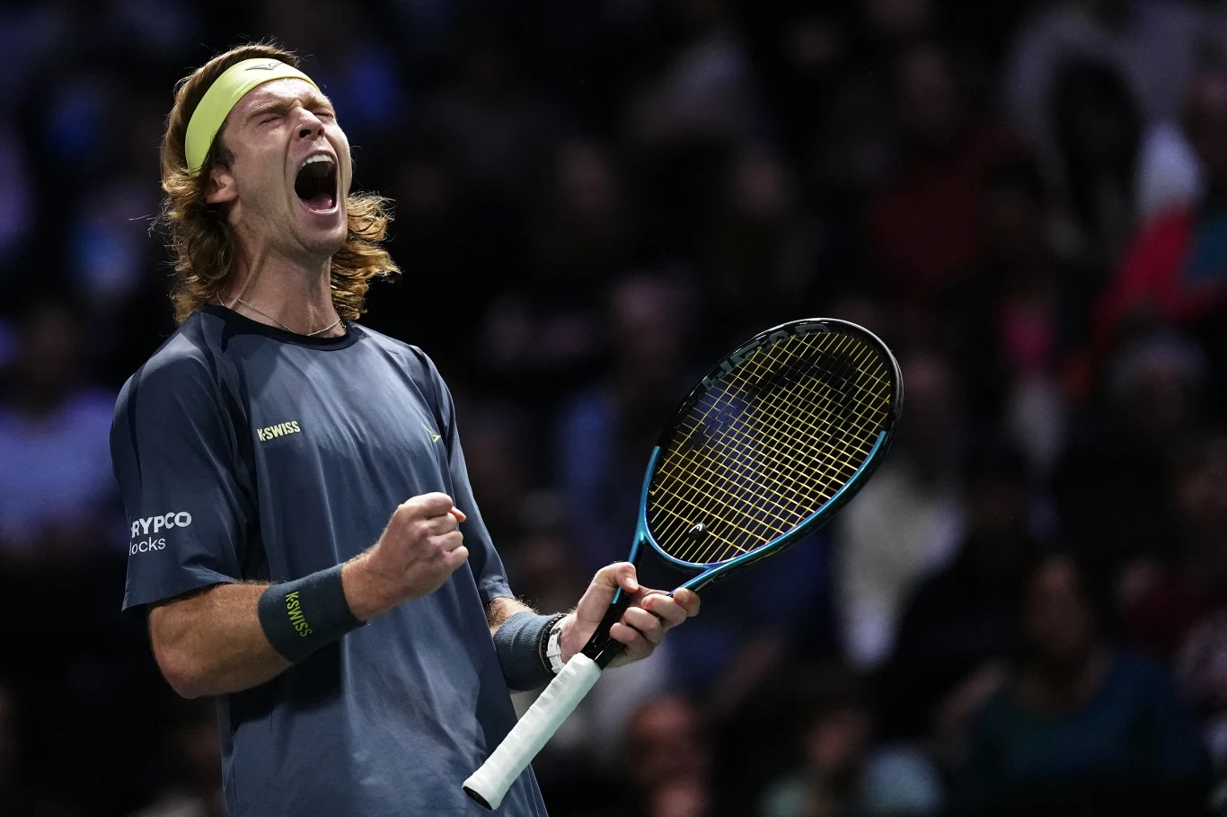 Młody mężczyzna z długimi blond włosami i opaską na głowie w sportowej koszulce trzyma rakietę tenisową, unosząc pięść i wyrażając silne emocje tryumfu lub radości w trakcie rozgrywki na korcie, w tle zamazana publiczność.