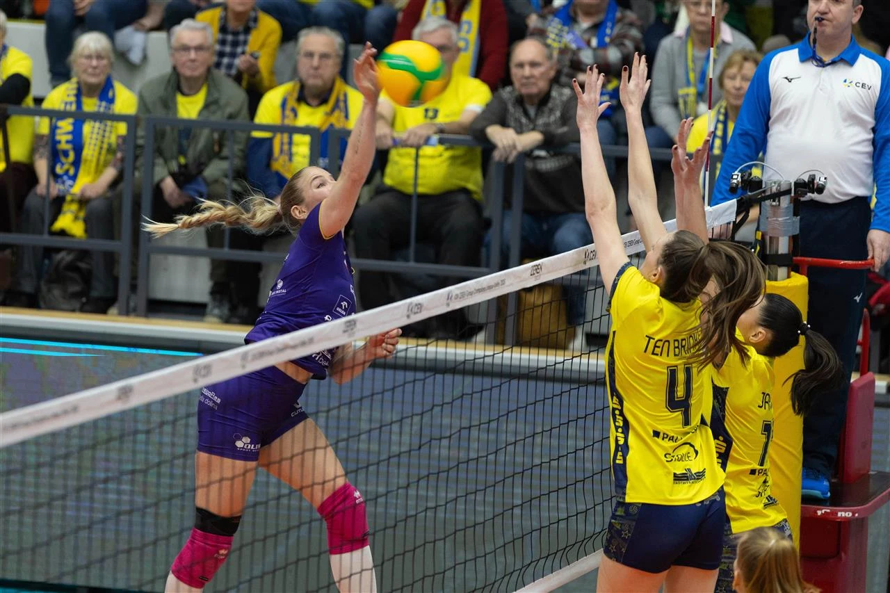 Akcja podczas meczu siatkówki kobiet, zawodniczka w fioletowym stroju atakuje piłkę ponad siatką, naprzeciwko dwie zawodniczki w żółtych strojach próbują zablokować uderzenie. W tle publiczność i sędzia na podwyższeniu.