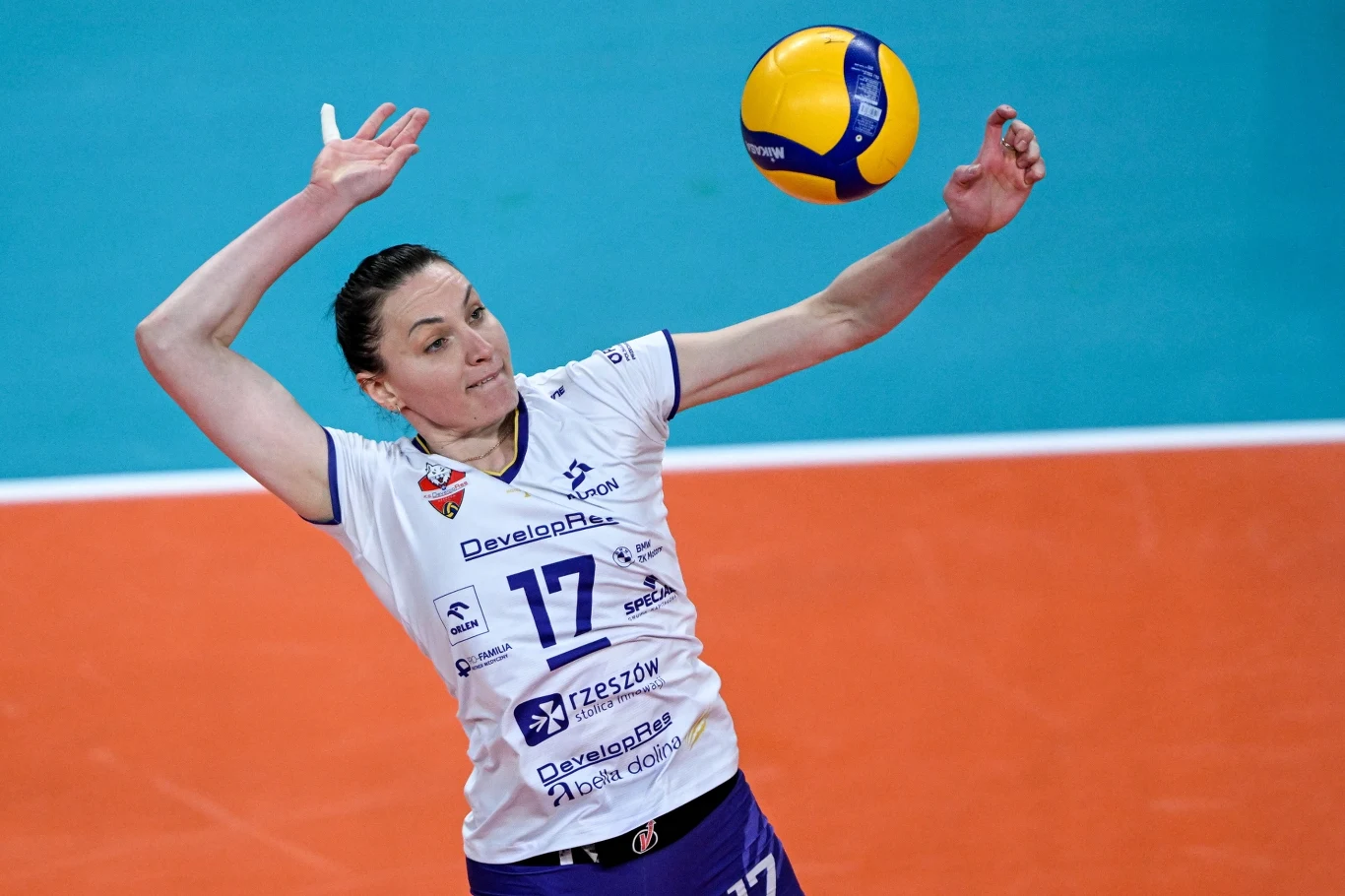 Siatkarka w białym stroju klubowym z numerem 17 wykonuje zagrywkę podczas meczu siatkówki, uniesiona ręka i intensywne skupienie w oczach, piłka znajduje się tuż przed dłonią zawodniczki, w tle widoczny fragment boiska o niebiesko-pomarańczowej nawierz...