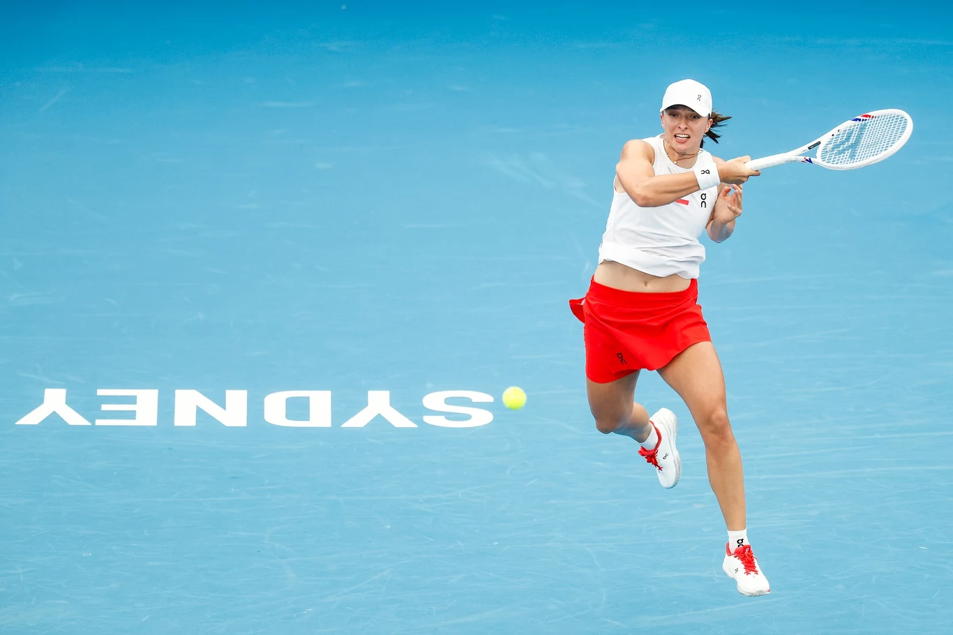 Iga Świątek Tenisistka w białej czapce i czerwonej spódnicy odbija piłkę na niebieskim korcie z widocznym napisem SYDNEY.