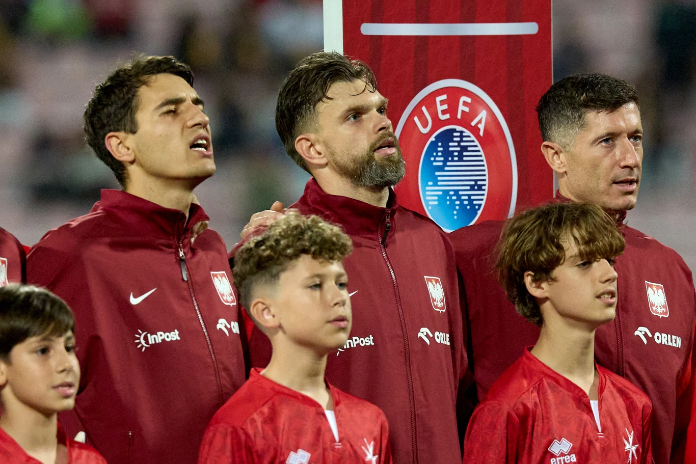 Jakub Kiwior, Bartłomiej Drągowski, Robert Lewandowski Grupa polskich piłkarzy narodowej reprezentacji oraz młodzi chłopcy w czerwonych strojach stoją przed tablicą z logo UEFA, wszyscy wpatrzeni przed siebie, część śpiewa hymn.