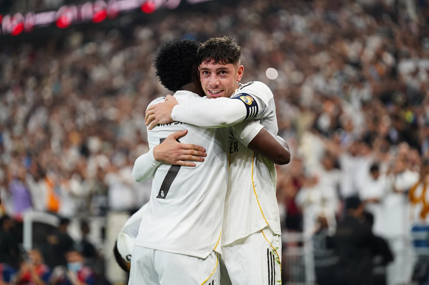 Vinicius i Federico Valverde Dwóch piłkarzy w białych strojach obejmuje się na boisku podczas meczu. W tle widać rozemocjonowanych kibiców na trybunach, którzy kibicują swojej drużynie.