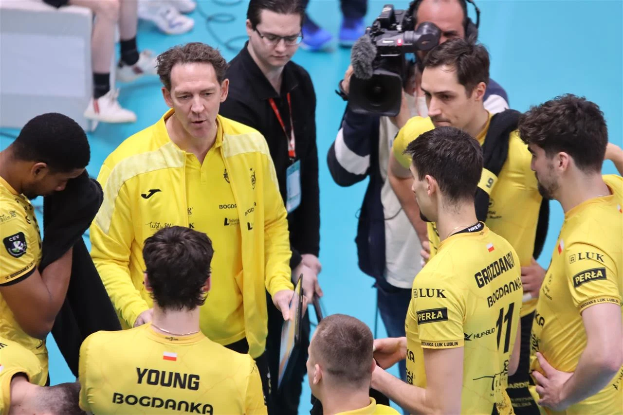 Trener drużyny siatkarskiej w żółtych strojach omawia strategię z zawodnikami podczas przerwy w meczu, obok stoi kamerzysta rejestrujący wydarzenie.