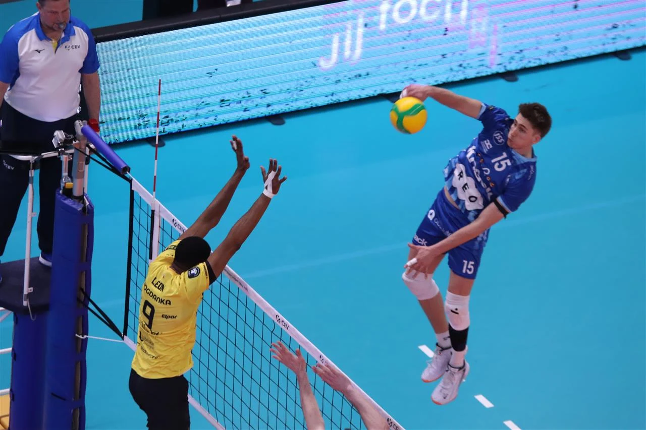 Siatkarz w niebieskim stroju wykonuje atak nad siatką, podczas gdy zawodnik w żółtej koszulce próbuje zablokować piłkę, obok nich stoi sędzia obserwujący przebieg akcji.