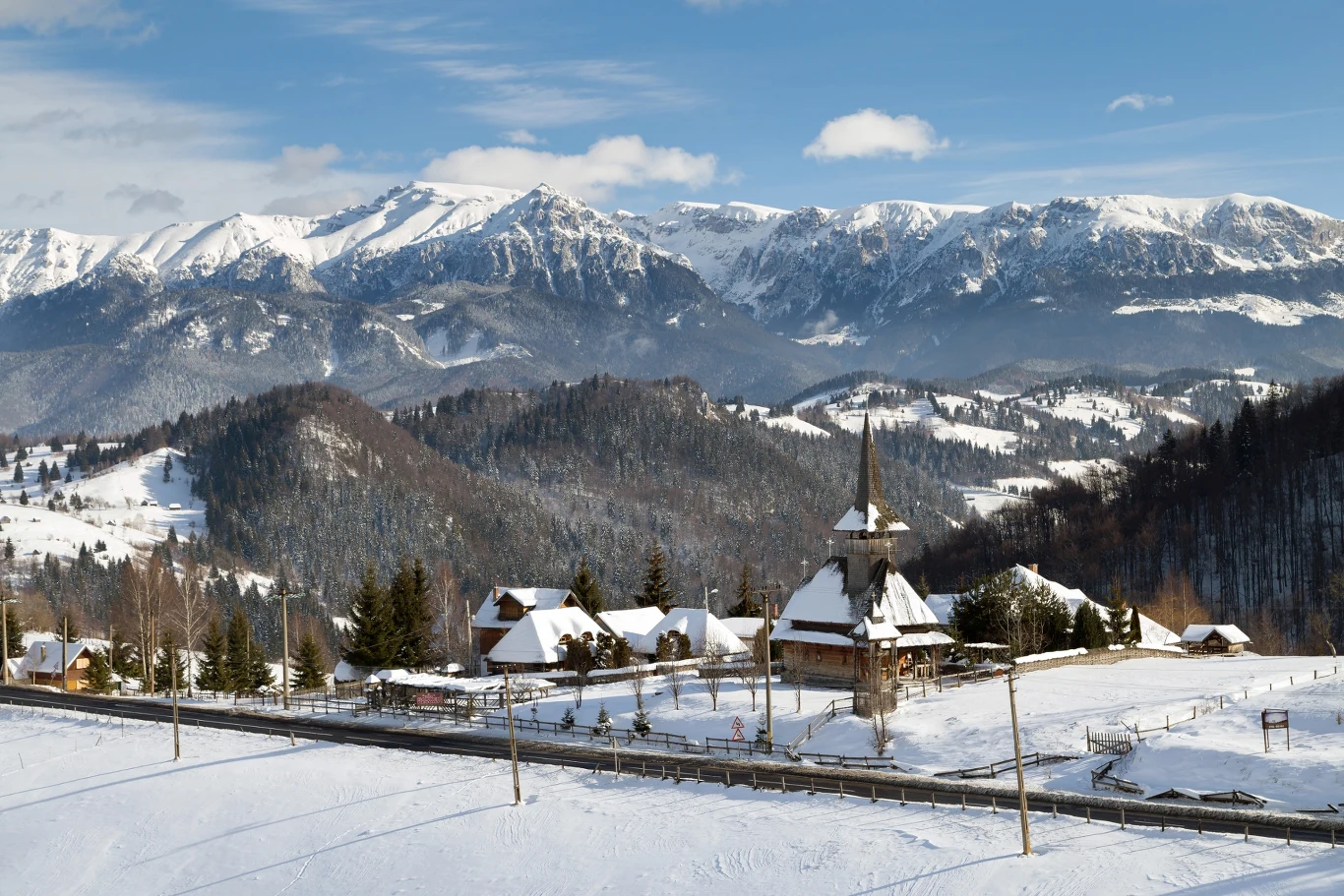Drewniany kościół z wysoką wieżą otoczony zabudowaniami i pokrytymi śniegiem łąkami, na tle majestatycznych, ośnieżonych gór oraz lasów.