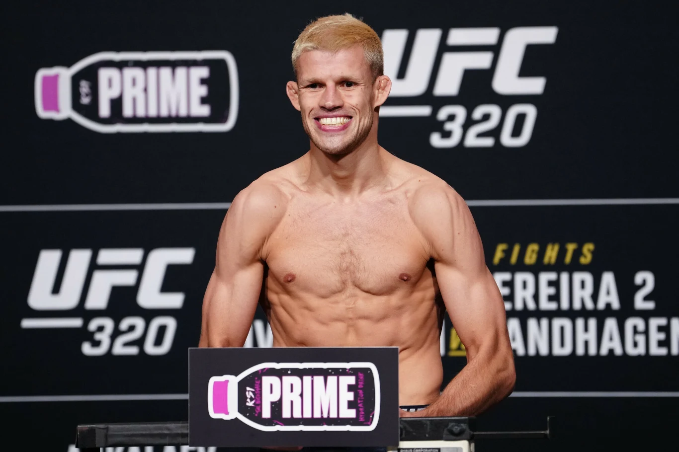 Jakub Wikłacz Uśmiechnięty mężczyzna o wysportowanej sylwetce stoi na wadze podczas oficjalnego ważenia przed galą UFC, w tle widoczne są logotypy sponsorów i numer gali.