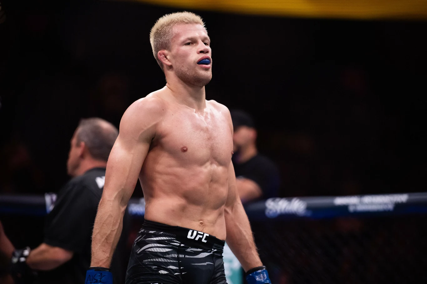 Jakub Wikłacz wraca do klatki UFC Zawodnik MMA stoi w oktagonie, ubrany w spodenki sportowe i ochraniacz na zęby, wyraża skupienie i gotowość do walki, w tle widoczni są sędziowie oraz fragmenty publiczności.