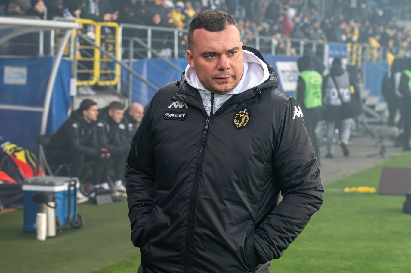 Mężczyzna ubrany w czarną kurtkę sportową z kapturem stoi na tle stadionu piłkarskiego, wokół niego widoczni są inni ludzie, trybuny oraz elementy infrastruktury boiska.