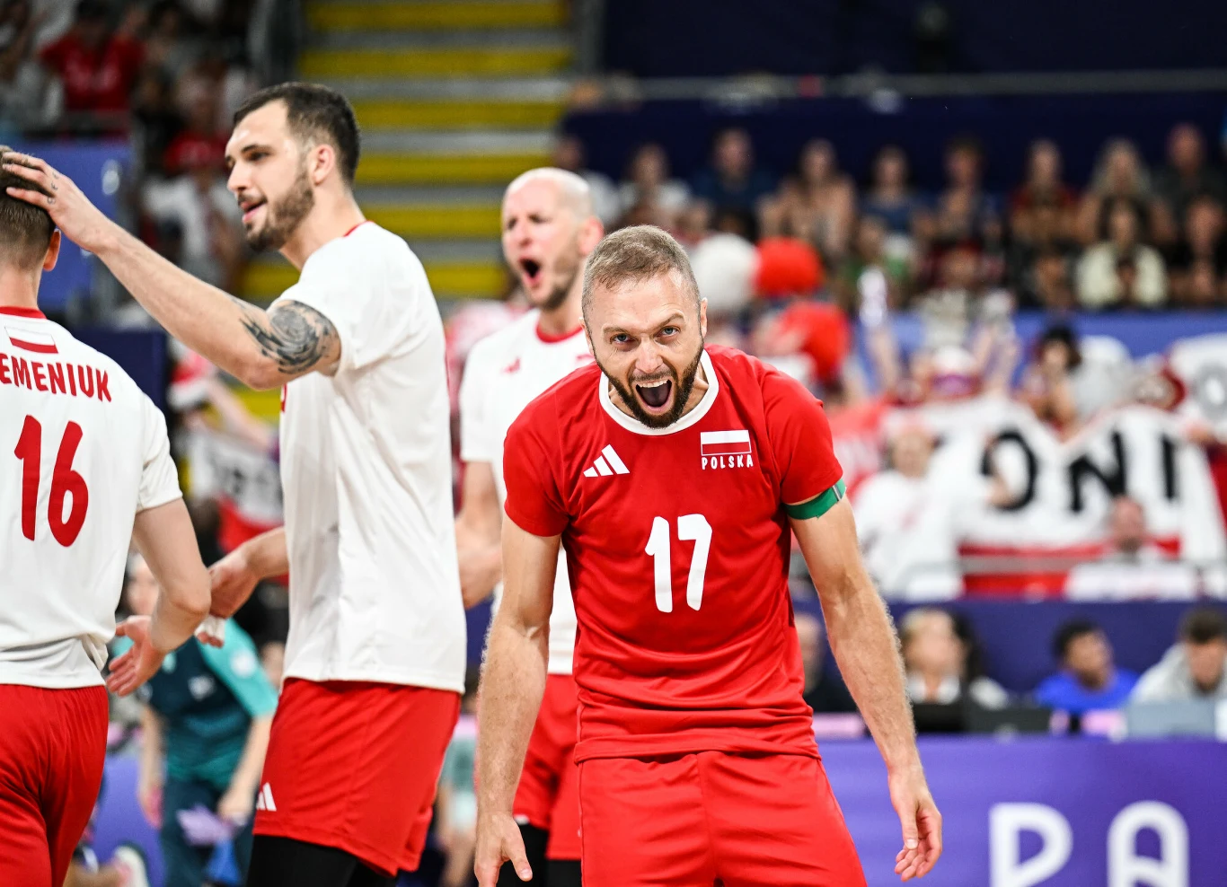 Polscy siatkarze podczas meczu na hali sportowej, zawodnik z numerem 17 w czerwonym stroju wyraża silne emocje, w tle widoczni pozostali członkowie drużyny oraz kibice na trybunach.