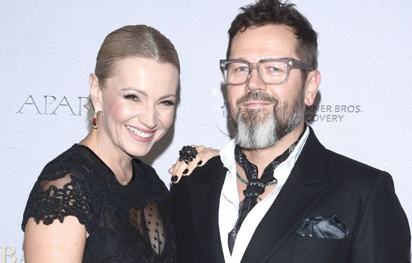 Dorota Szelągowska i Wojciech Wysocki Dwie elegancko ubrane osoby pozujące razem na tle jasnej ściany z nadrukowanymi nazwami sponsorów lub firm. Kobieta w czarnej koronkowej sukni uśmiecha się szeroko, mężczyzna w ciemnym garniturze, z brodą i okulary, ma apaszkę i również uśmiecha się le...