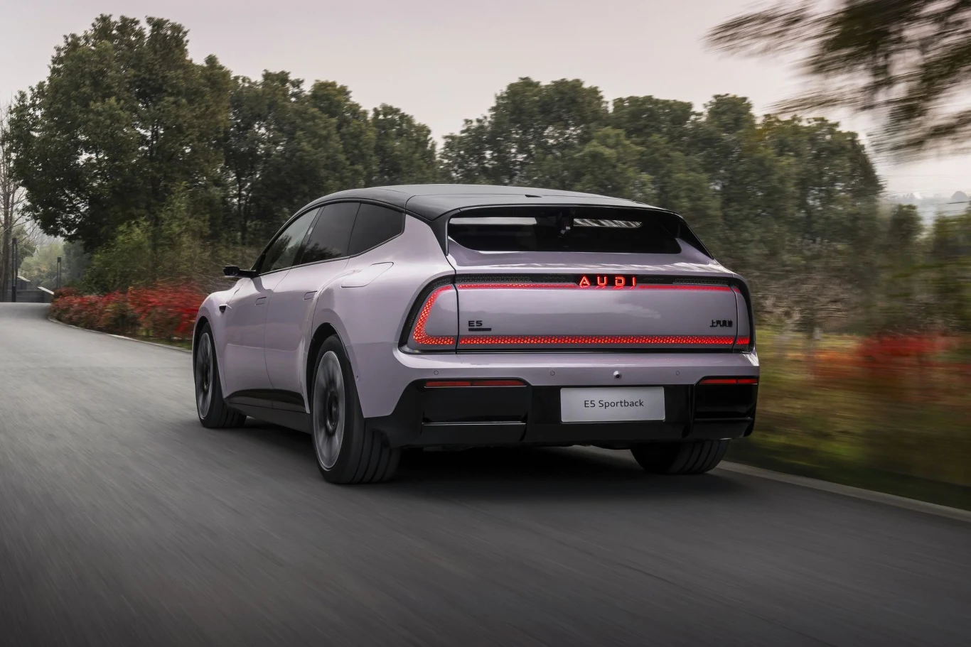 Nowoczesny samochód elektryczny Audi w jasnym kolorze porusza się po asfaltowej drodze otoczonej drzewami, widok zza auta eksponuje charakterystyczne światła tylne i dynamiczną sylwetkę.