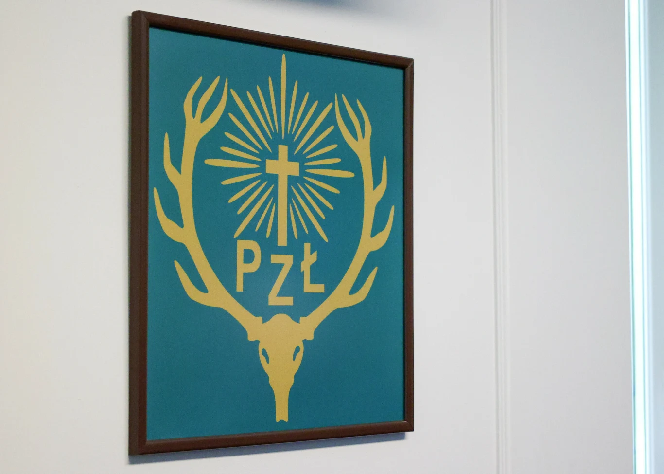Herb przedstawiający złotą czaszkę jelenia z porożem, krzyżem i promieniami światła oraz literami PZŁ na niebieskim tle, w drewnianej ramie, zawieszony na jasnej ścianie.