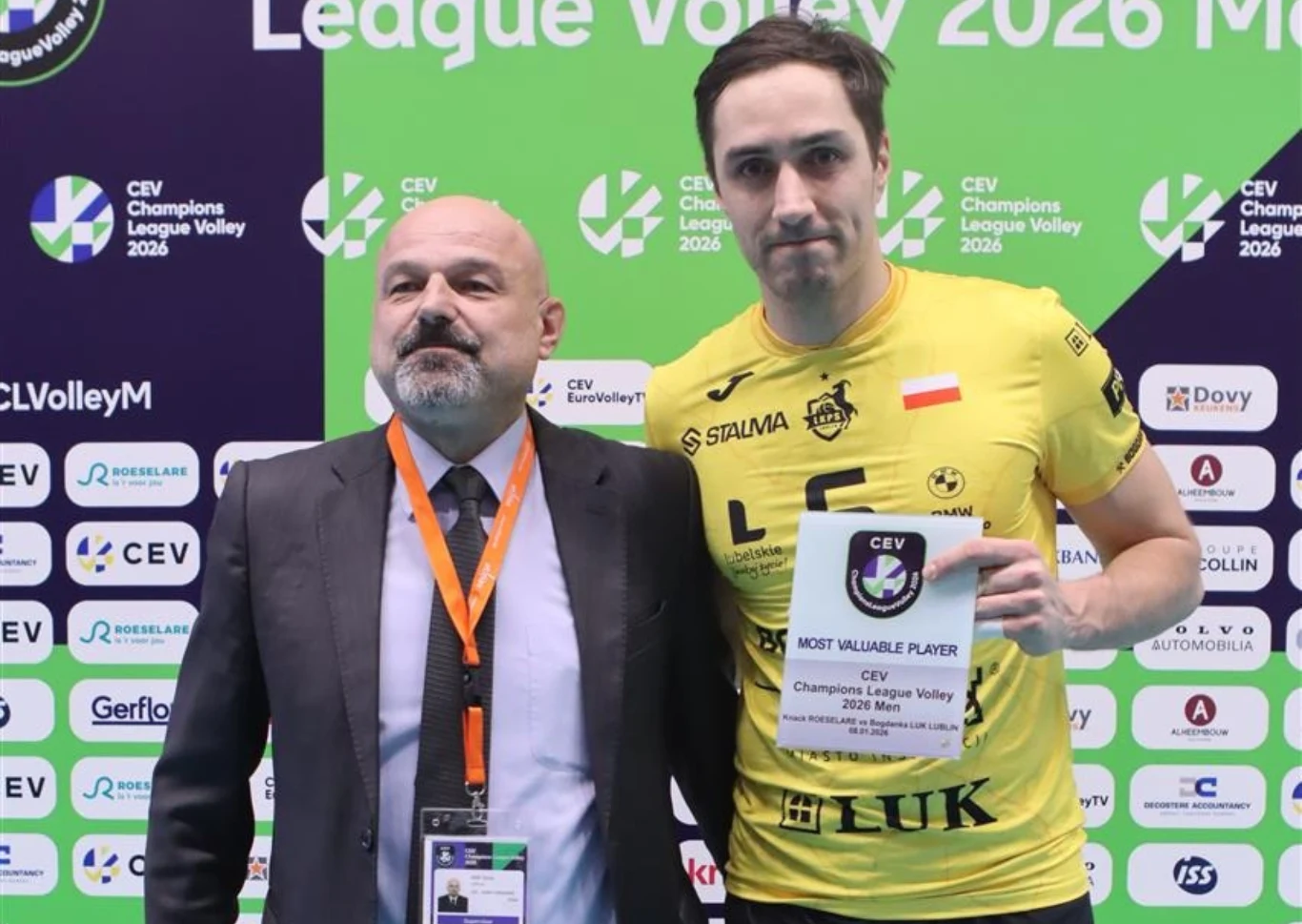 Dwóch mężczyzn stojących razem na tle ścianki sponsorsko-turniejowej podczas wydarzenia siatkarskiego, jeden z nich ubrany w żółtą koszulkę sportową trzyma tabliczkę z napisem Most Valuable Player, drugi ma na sobie garnitur oraz zawieszkę z identyfika...