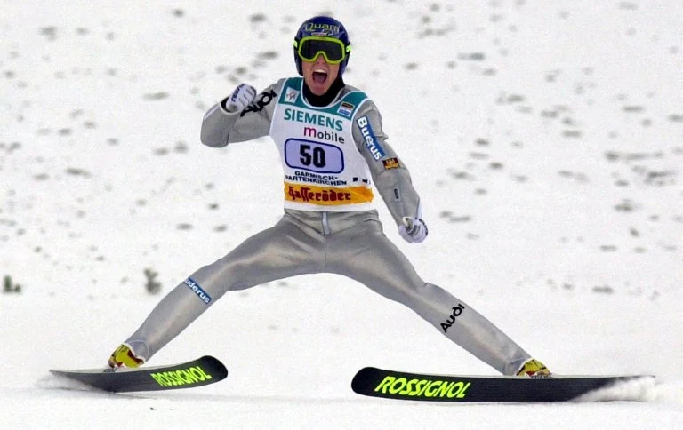 Sportowiec w stroju do skoków narciarskich wykonuje dynamiczny manewr po wylądowaniu na śnieżnym stoku, szeroko rozstawiając narty i świętując swój wynik emocjonalnym gestem.