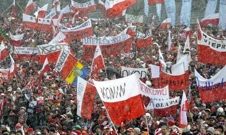 Tłum ludzi ubranych na biało-czerwono, trzymający liczne polskie flagi i transparenty, otoczony opadami śniegu, atmosfera podniosła, wiele osób świętuje i wspiera wspólną sprawę.