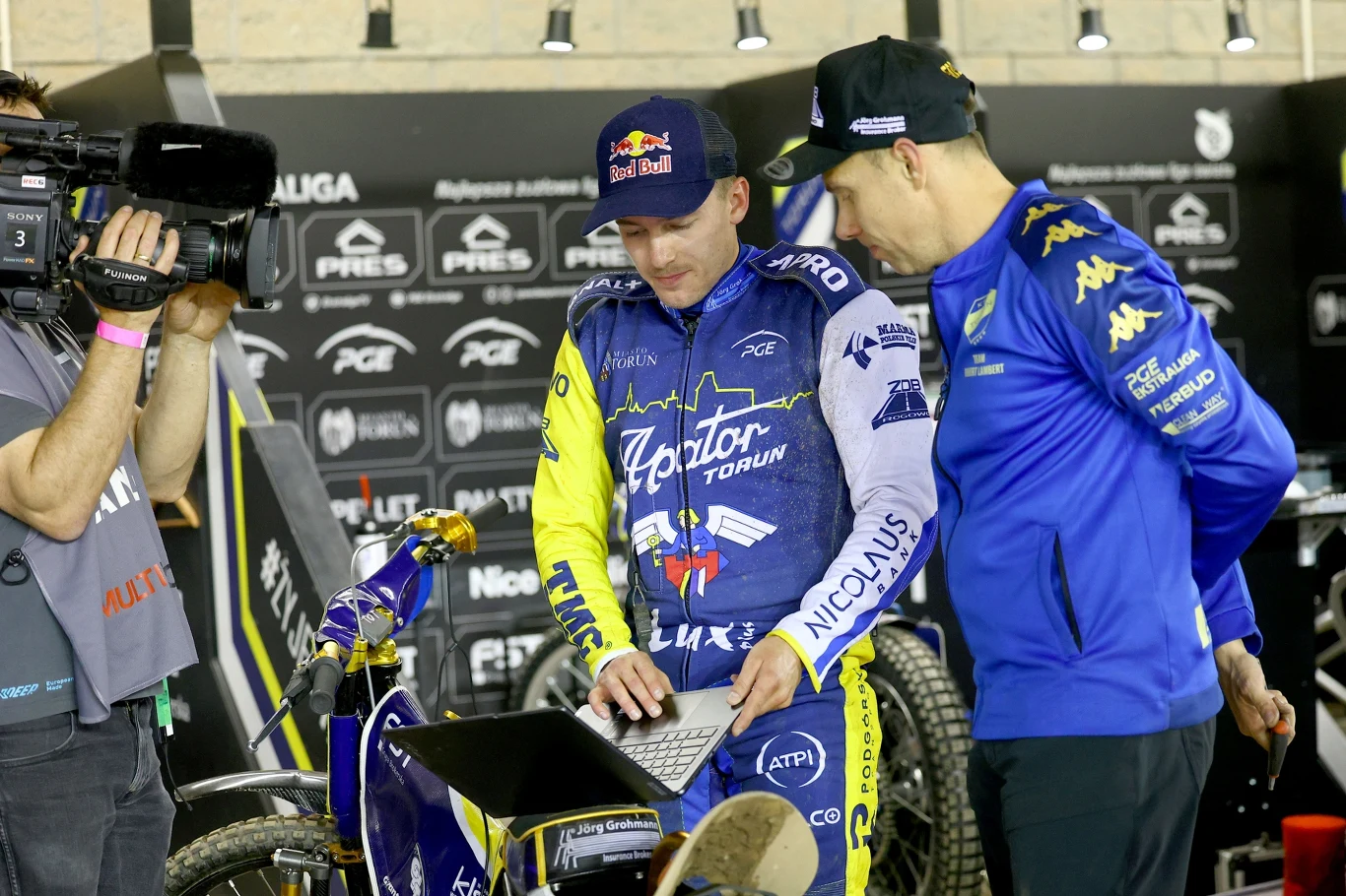 Dwóch mężczyzn ubranych w stroje sportowe z logo zespołów żużlowych analizuje dane na laptopie przy motocyklu żużlowym, obok nich kamerzysta nagrywa materiał w wyraźnie sportowym otoczeniu, tłem są banery sponsorów.