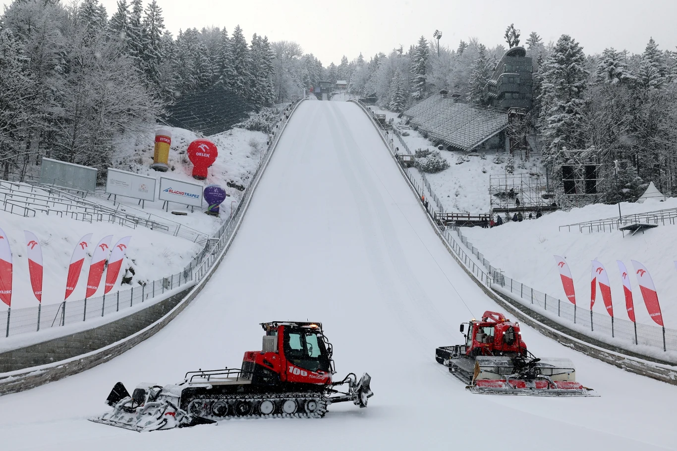 Dwie ratraki przygotowujące skocznię narciarską w zimowym krajobrazie, otoczoną świerkami oraz trybunami po bokach stoku. Śnieg pokrywa całą infrastrukturę sportową i drzewa, dodając zimowej atmosfery.