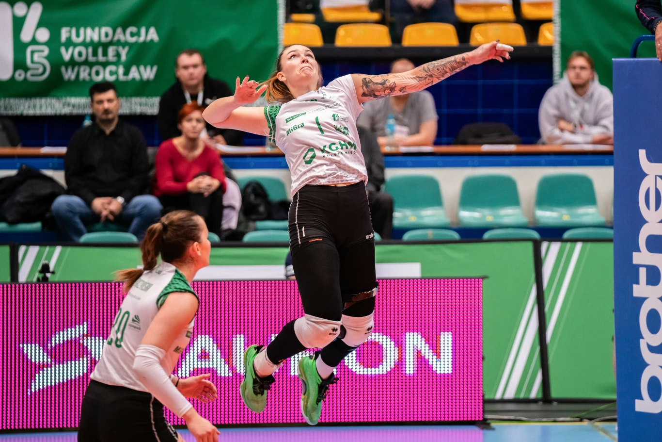 Zawodniczka w trakcie wyskoku do ataku podczas meczu siatkówki, w tle widoczni kibice na trybunach oraz banery sponsorskie. Obok niej znajduje się druga siatkarka przygotowująca się do gry.