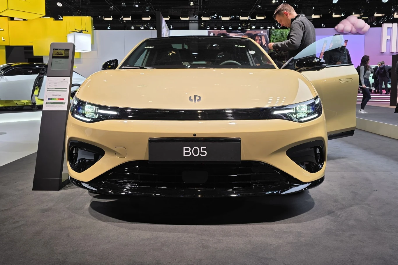 Beżowy samochód elektryczny BO5 ustawiony na stoisku targowym w jasnej hali wystawowej, z otwartymi drzwiami po stronie kierowcy i mężczyzną oglądającym wnętrze pojazdu.