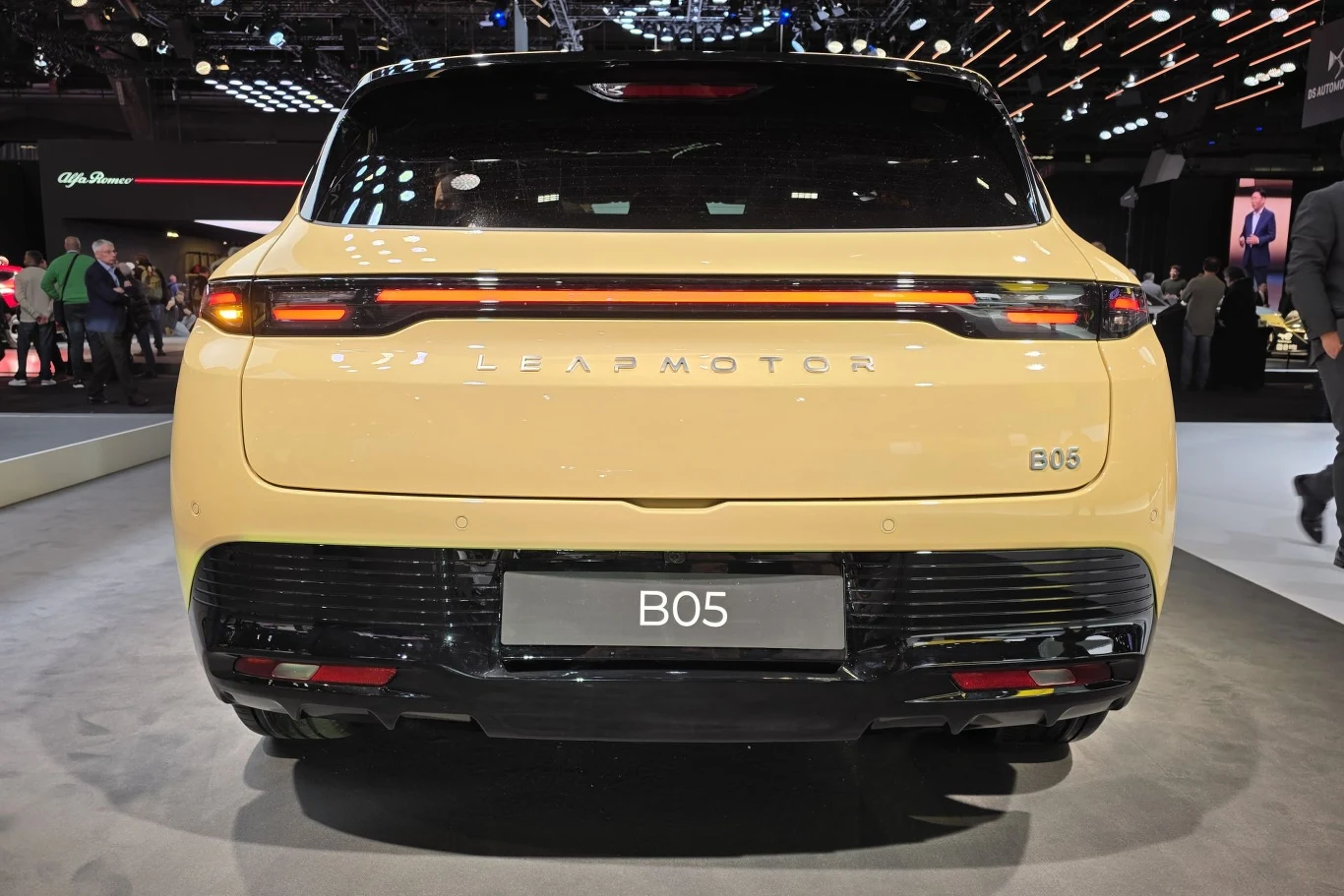 Tylna część samochodu osobowego marki Leapmotor B05 w kolorze żółtym, widoczne nowoczesne światła tylne, napis z nazwą producenta oraz modelu, auto wystawione na hali targowej, w tle osoby i elementy ekspozycji.