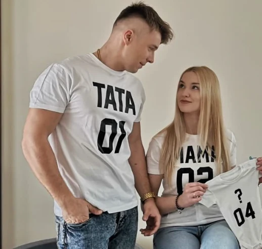 Para młodych ludzi uśmiecha się do siebie, ubrani w białe koszulki z napisami „TATA 01” i „MAMA 02”. Kobieta trzyma w ręku malutką białą koszulkę z napisem „? 04”, co sugeruje oczekiwanie na dziecko.