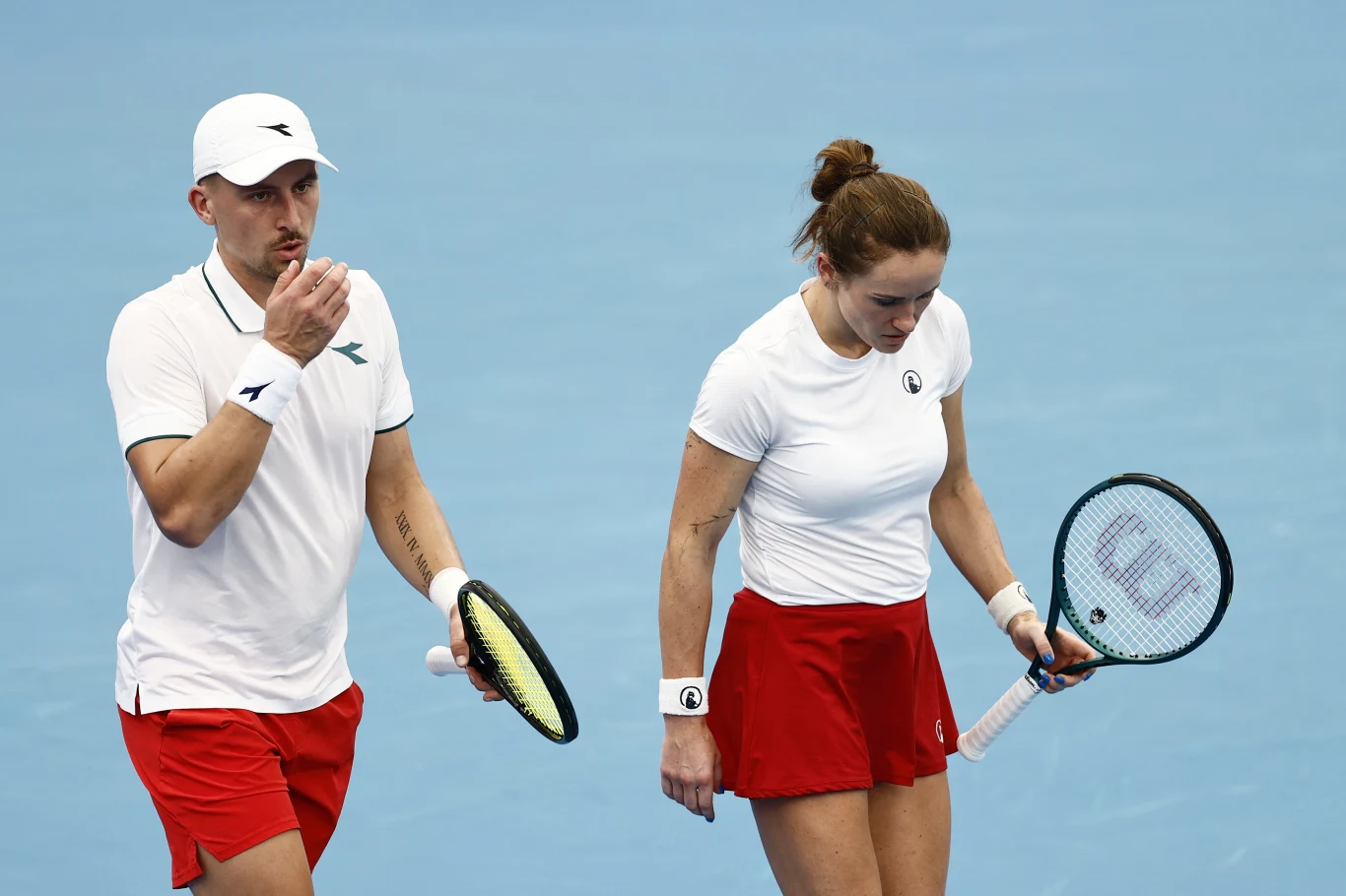 Dwóch tenisistów na niebieskim korcie, ubrani w białe koszulki i czerwone spodenki, trzymają rakiety w rękach. Kobieta patrzy w dół, mężczyzna trzyma piłkę tenisową i wykonuje gest ustami.