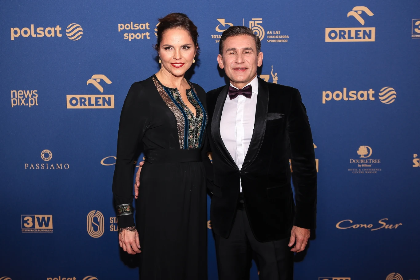 Robert Korzeniowski z żoną na Gali Mistrzów Sportu, 2022 Dwie elegancko ubrane osoby stoją na tle ścianki z logotypami sponsorów, kobieta w długiej czarnej sukni z ozdobnym dekoltem, mężczyzna w czarnym garniturze i muchą, uśmiechają się i pozują do zdjęcia.