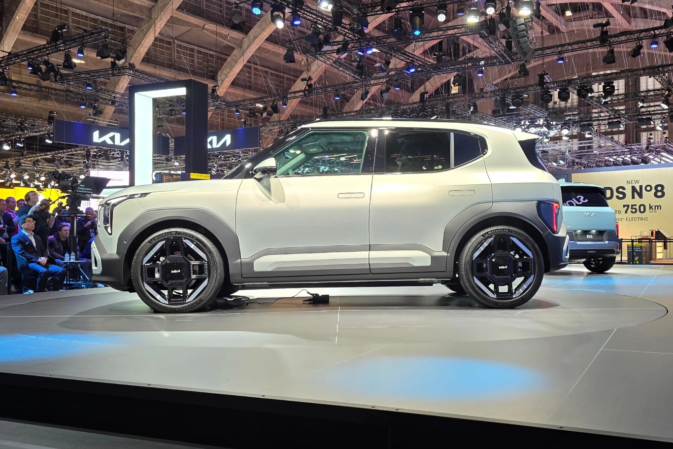 Nowoczesny biały samochód terenowy eksponowany na oświetlonej scenie targów motoryzacyjnych, w tle widoczne stoiska innych marek oraz elementy infrastruktury hali wystawowej.