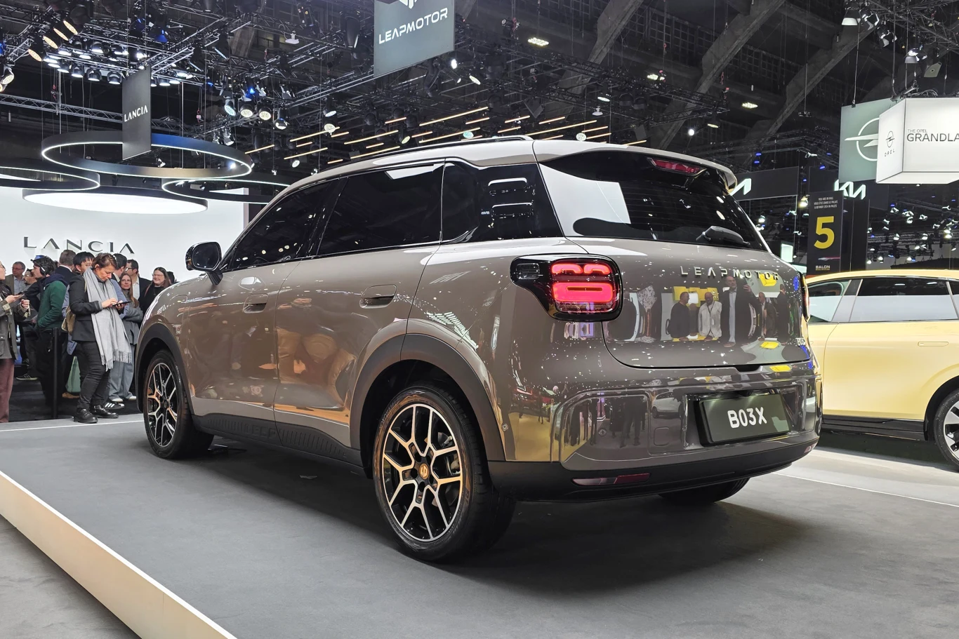 Nowoczesny, szary SUV marki Leapmotor B03X ustawiony na podwyższeniu na targach motoryzacyjnych. W tle widoczna grupa ludzi oraz inne samochody w jasnym oświetleniu hali wystawowej.