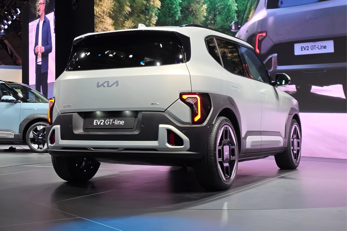 Nowoczesny srebrny samochód elektryczny Kia EV2 GT-line ustawiony na ekspozycji targowej z widocznym oświetleniem LED oraz charakterystycznymi, geometrycznymi felgami. W tle fragment innego auta i ekran prezentacyjny.