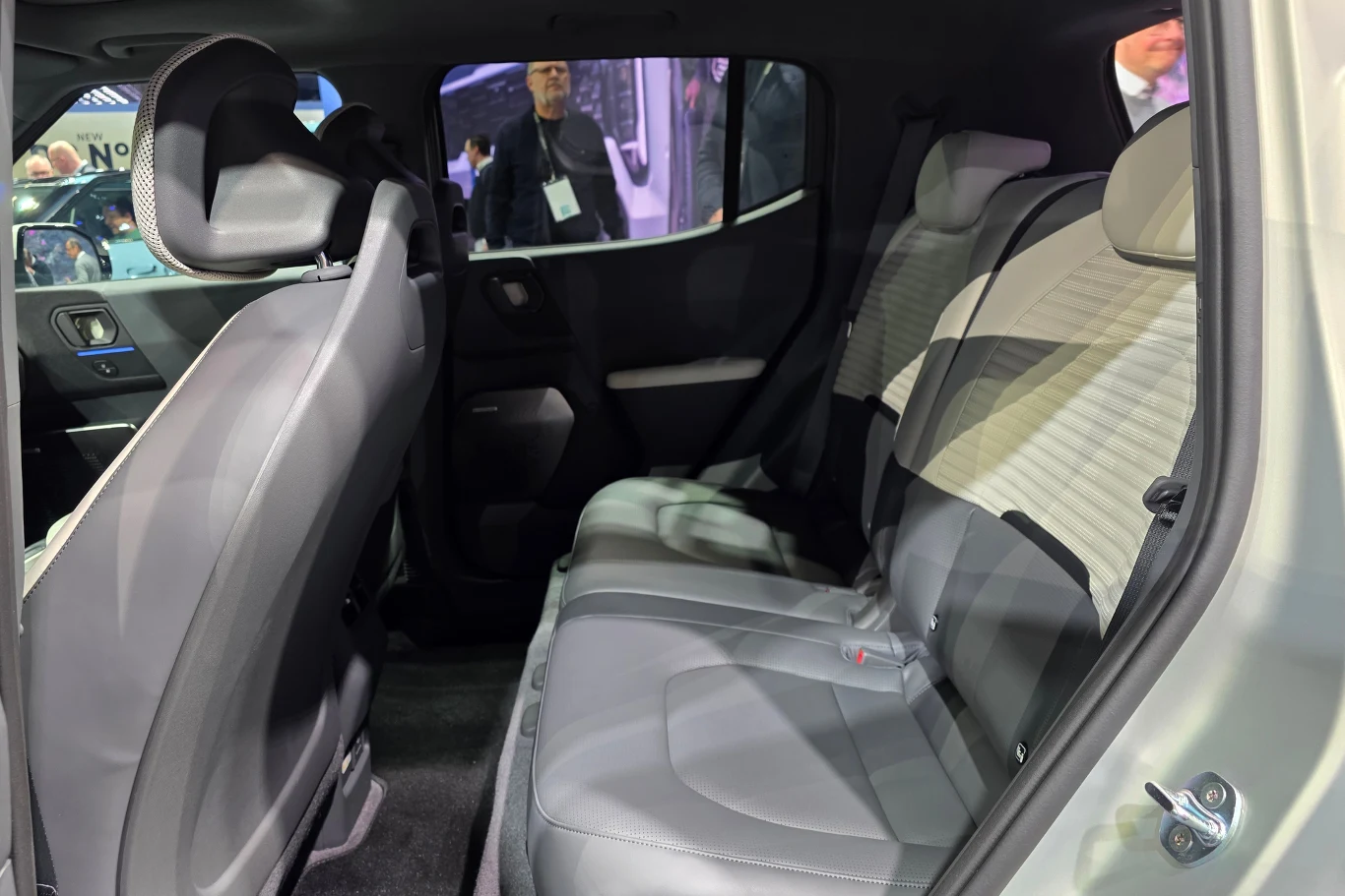 Wnętrze samochodu osobowego z widokiem na tylne siedzenia, drzwi i przestrzeń pasażerską, wykończone jasnoszarą tapicerką. W tle przez okno widoczne sylwetki ludzi stojących na zewnątrz pojazdu.