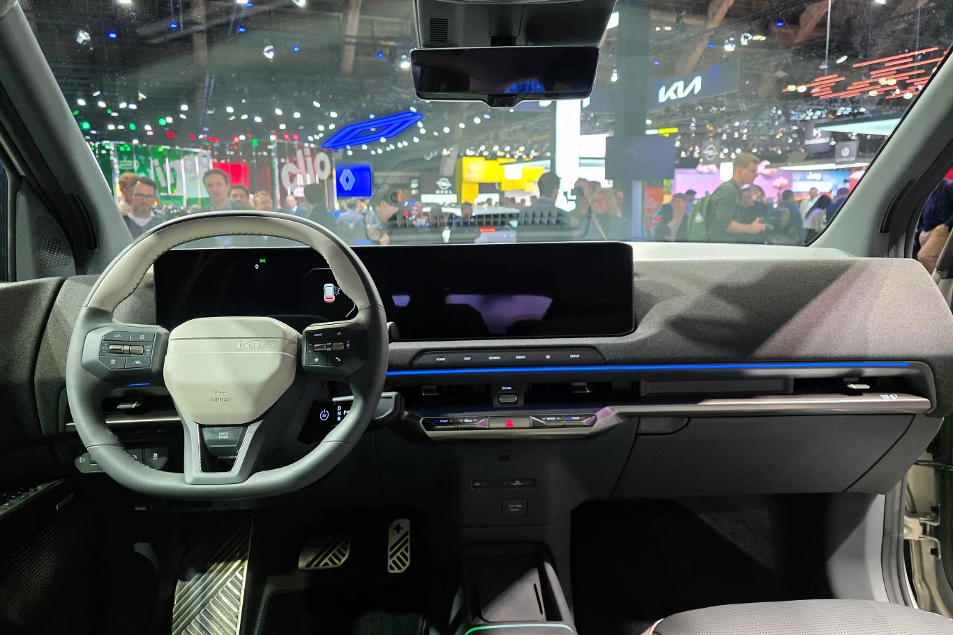 Nowoczesna deska rozdzielcza samochodu elektrycznego z szerokim cyfrowym wyświetlaczem, wielofunkcyjną kierownicą oraz minimalistycznym wykończeniem kokpitu. W tle widoczne rozświetlone stanowiska targowe i osoby zwiedzające.