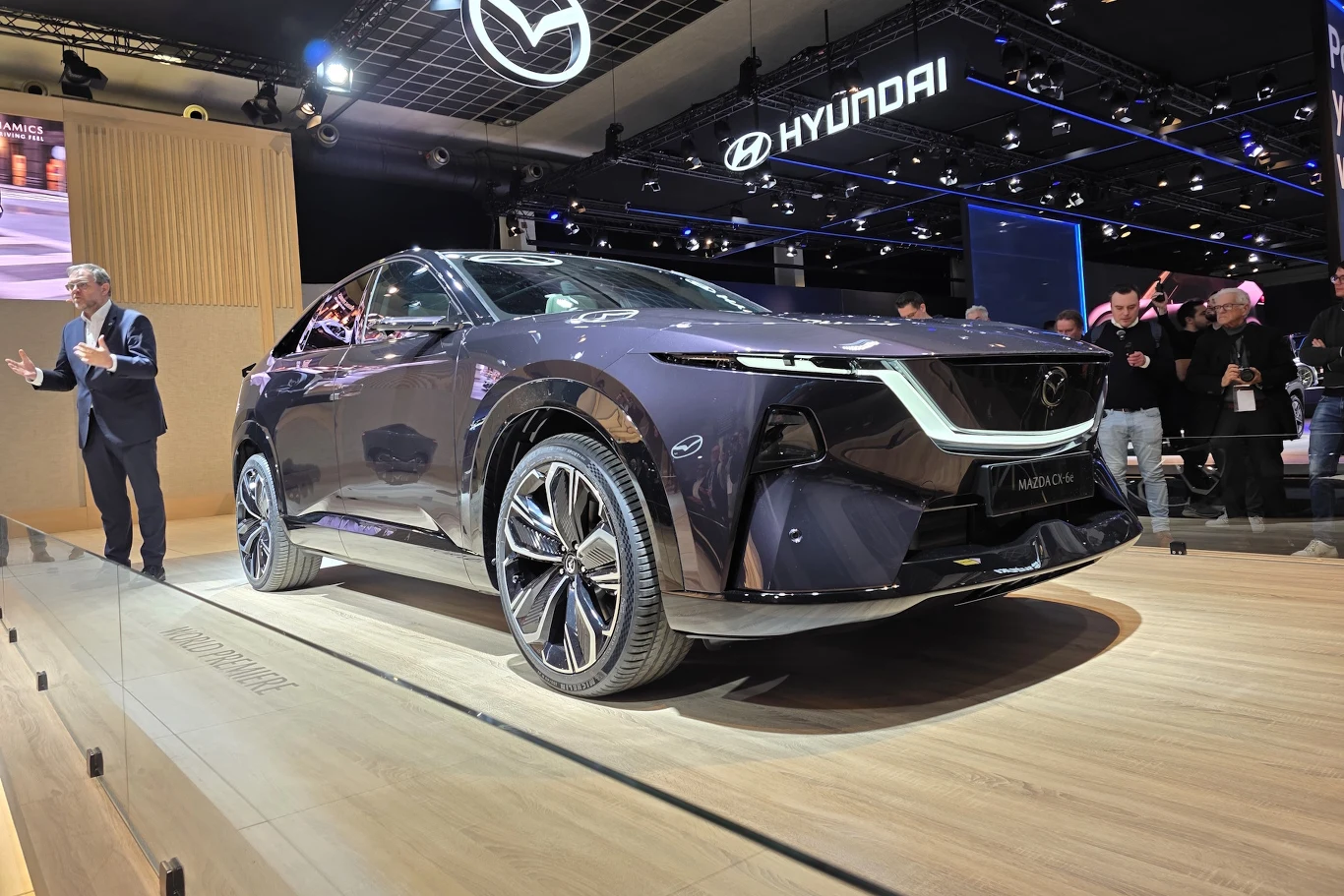 Mazda CX-6e Czarny nowoczesny samochód koncepcyjny Mazda eksponowany na podwyższeniu w hali targowej, w tle widoczne logo Mazda i Hyundai, a obok samochodu stoi mężczyzna przemawiający do publiczności.