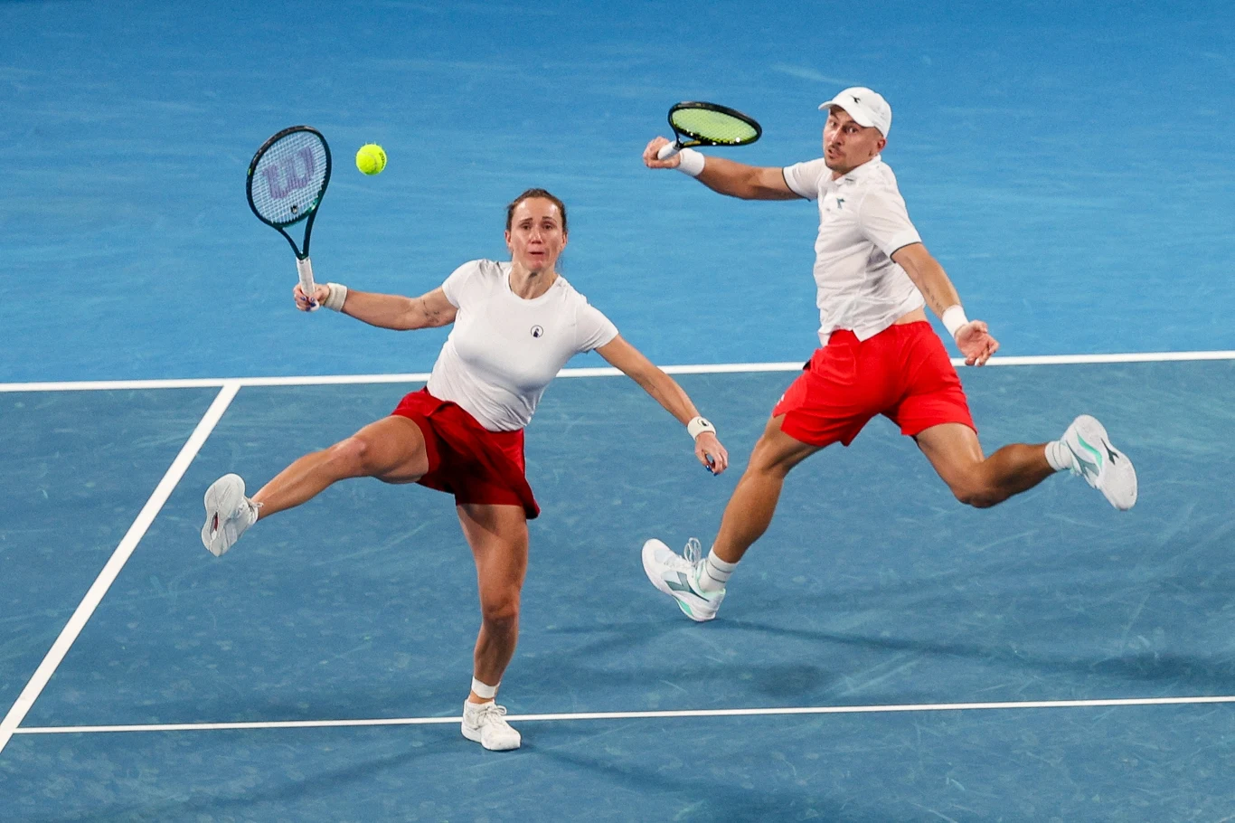Dwoje tenisistów w czerwono-białych strojach dynamicznie wykonuje uderzenie rakietami, skupieni na piłce tenisowej znajdującej się w powietrzu nad niebieskim kortem.