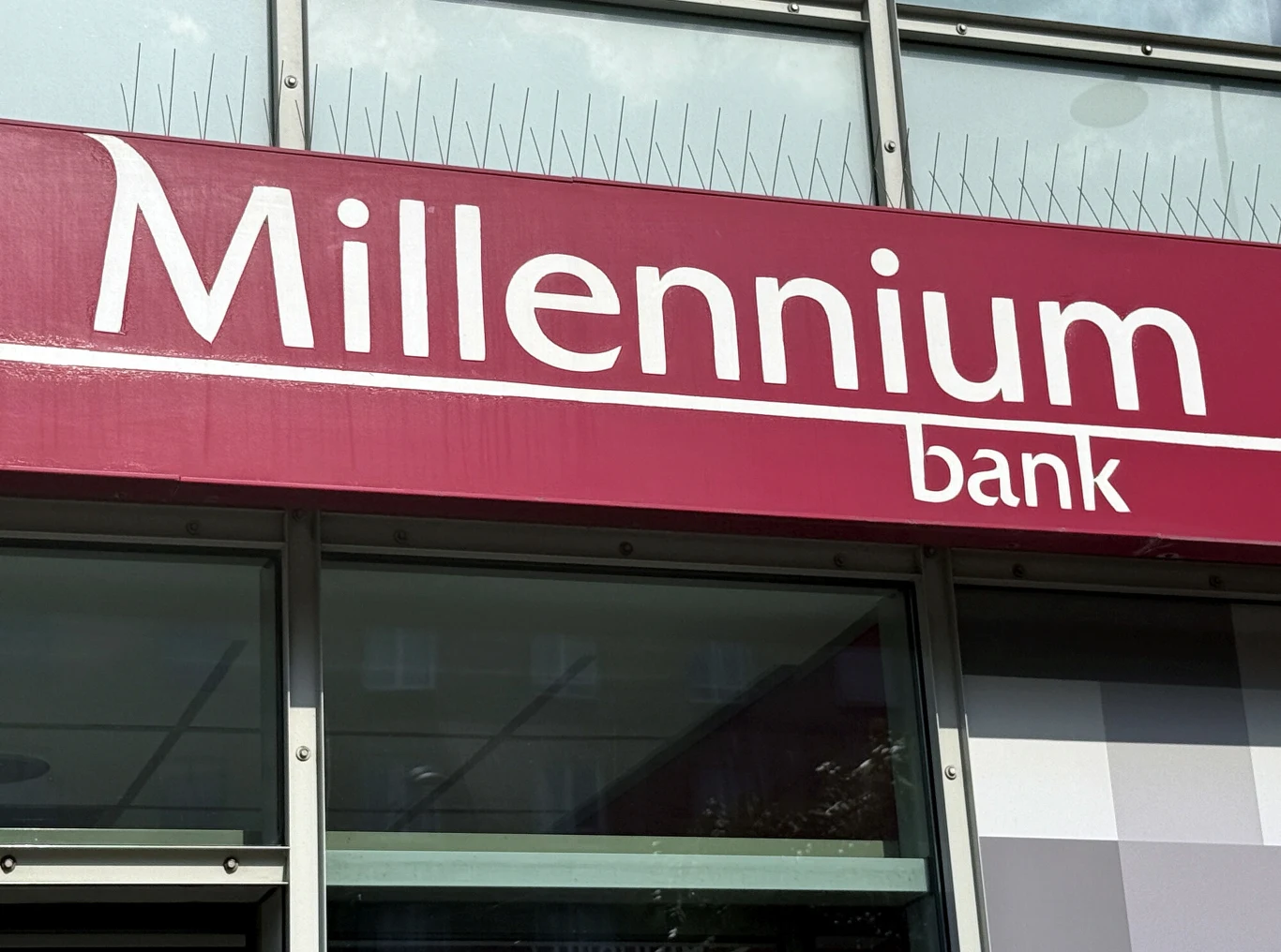 Szyld banku Millennium na bordowym tle, zamontowany na elewacji budynku, widoczne napisy w białym kolorze oraz elementy okna pod szyldem.