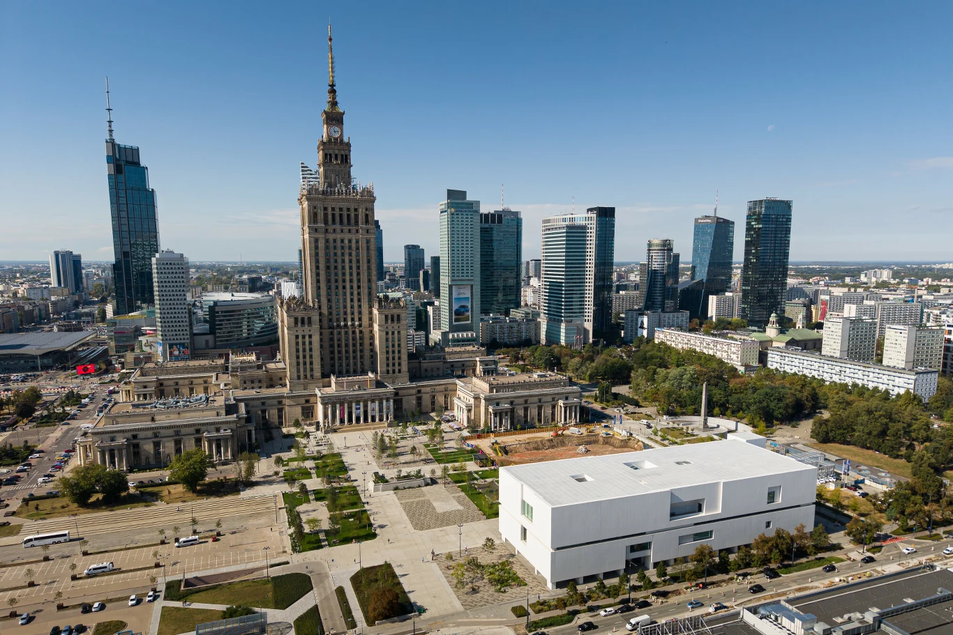 Centrum Warszawy z dominującym Pałacem Kultury i Nauki otoczonym nowoczesnymi wieżowcami i biurowcami, na pierwszym planie biały budynek Muzeum Sztuki Nowoczesnej, rozległe place i alejki spacerowe.