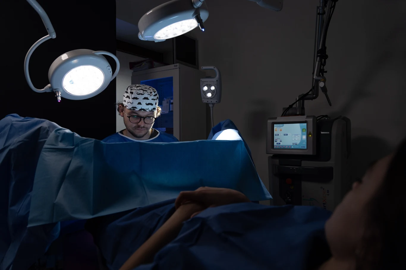 Chirurg w sali operacyjnej przeprowadza zabieg pod jasnym oświetleniem zabiegowym. W tle widoczny monitor z parametrami pacjenta oraz sprzęt medyczny. Pacjentka leży na stole operacyjnym, przykryta niebieskim materiałem.