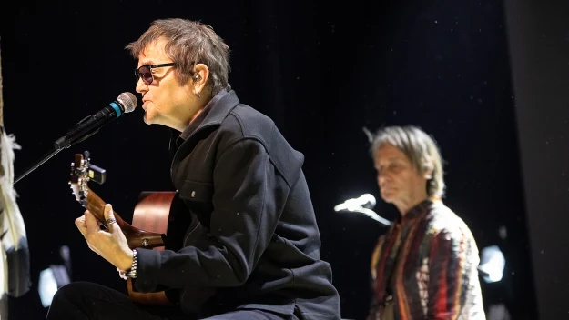 Mężczyzna w okularach śpiewający do mikrofonu i grający na gitarze akustycznej, w tle siedzi druga osoba w kolorowej koszuli, scena oświetlona światłami scenicznymi.