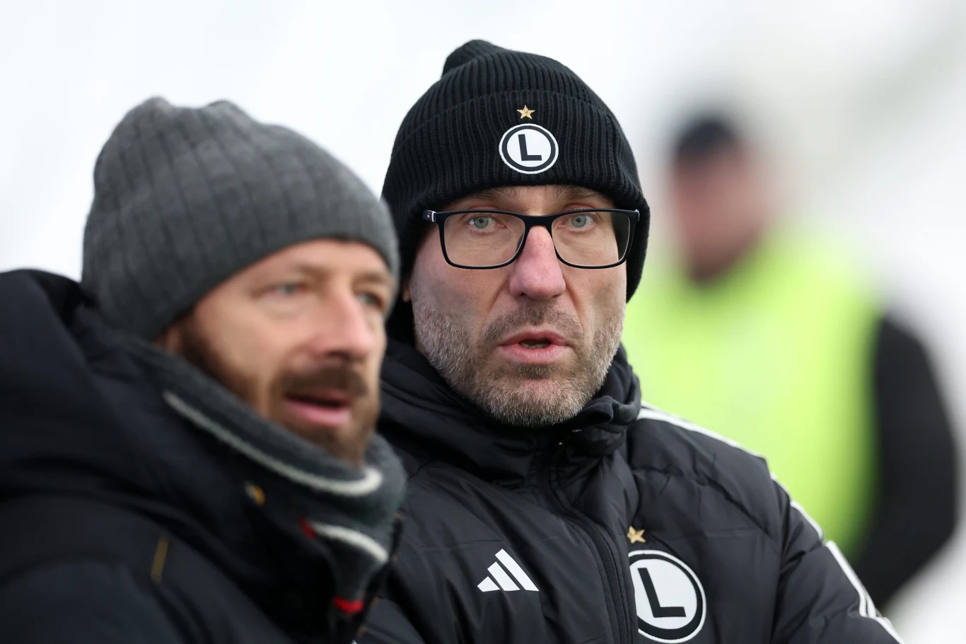 Marek Papszun Dwie osoby ubrane w zimowe kurtki, czapki i szaliki z logo klubu sportowego Legia Warszawa stoją na zewnątrz, w tle postać w jasnozielonej kurtce widoczna nieostro.