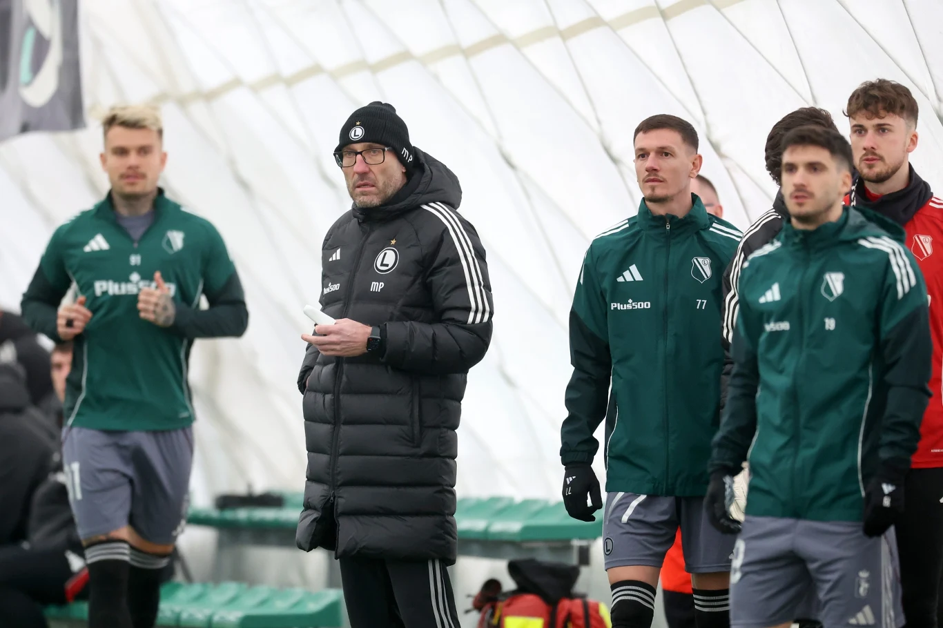 Legia Warszawa Grupa trenerów i zawodników w zielonych i czarnych strojach sportowych stoi pod białym namiotem, główny trener ubrany w czarną kurtkę puchową i czapkę koncentracją obserwuje boisko, obok nich zielone ławki dla rezerwowych.