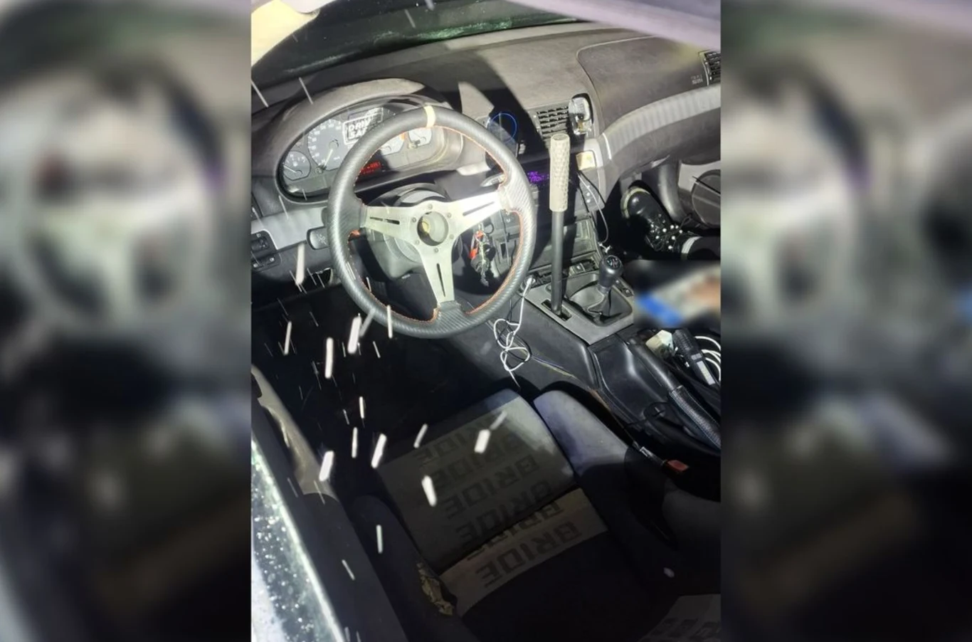 Wnętrze sportowego samochodu z widoczną kierownicą, dźwignią zmiany biegów oraz kubełkowym fotelem BRIDE. Część kokpitu w zbliżeniu, z przewodami i akcesoriami elektronicznymi. Na siedzeniu przednim odblaskowe elementy świetlne.