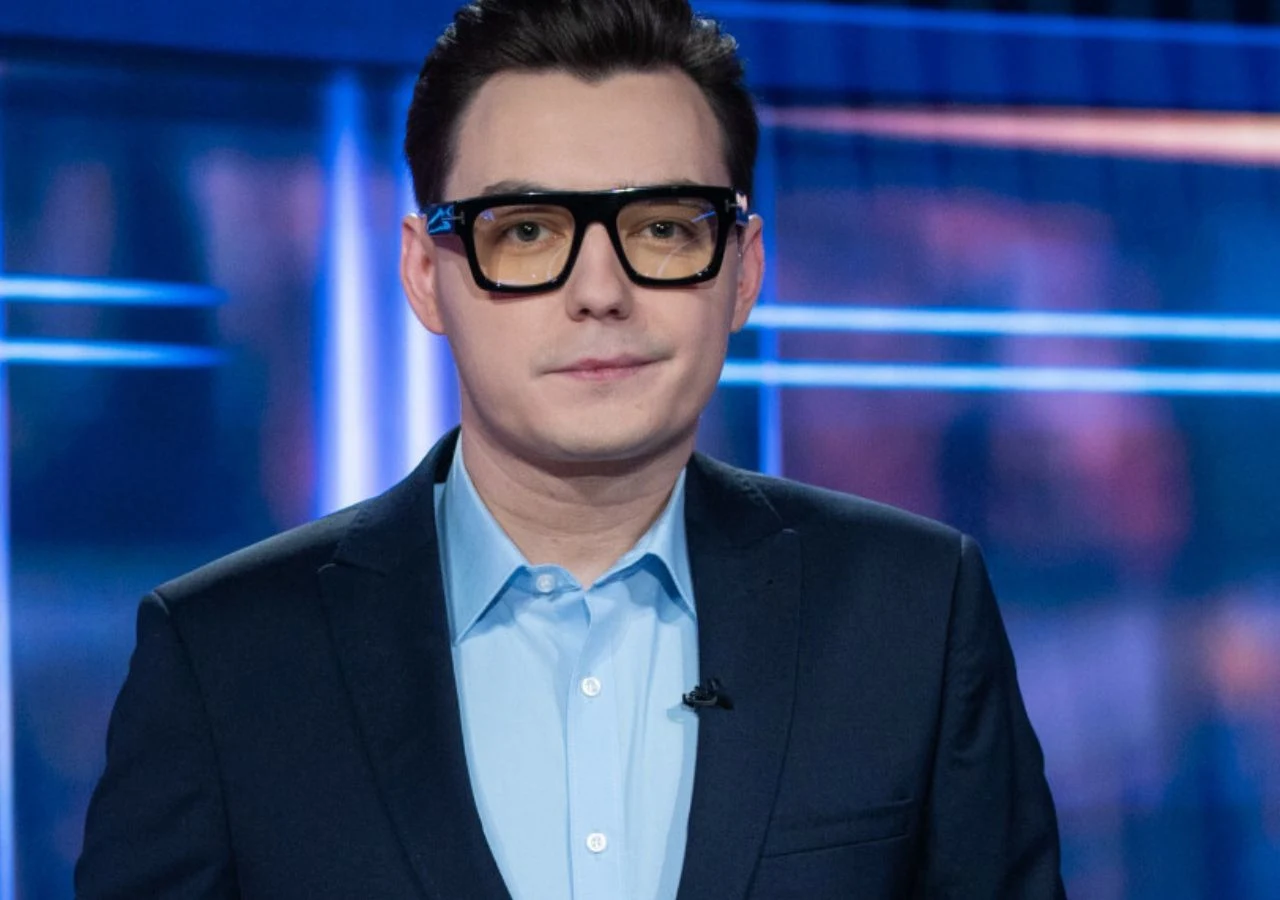 Bartosz Cebeńko, "Teleexpress" Mężczyzna w ciemnych okularach i garniturze stoi na tle niebiesko-fioletowego studia telewizyjnego, patrząc bezpośrednio w obiektyw.