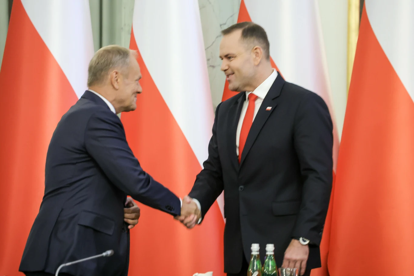Premier Donald Tusk zabrał głos po spotkaniu z prezydentem Karolem Nawrockim (zdj. ilustracyjne) Dwóch mężczyzn w garniturach podaje sobie ręce na tle kilku biało-czerwonych flag Polski, w otoczeniu mikrofonów i butelek z wodą, co sugeruje oficjalne spotkanie.