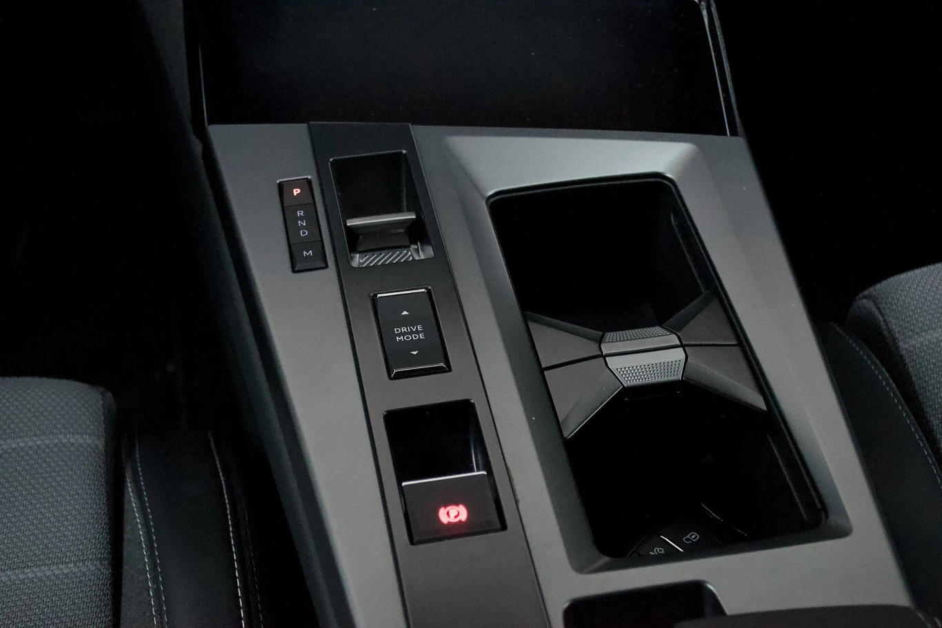 Konsola środkowa samochodu z widocznymi przyciskami do sterowania różnymi funkcjami, miejscem na napoje oraz tapicerką siedzenia kierowcy i pasażera.