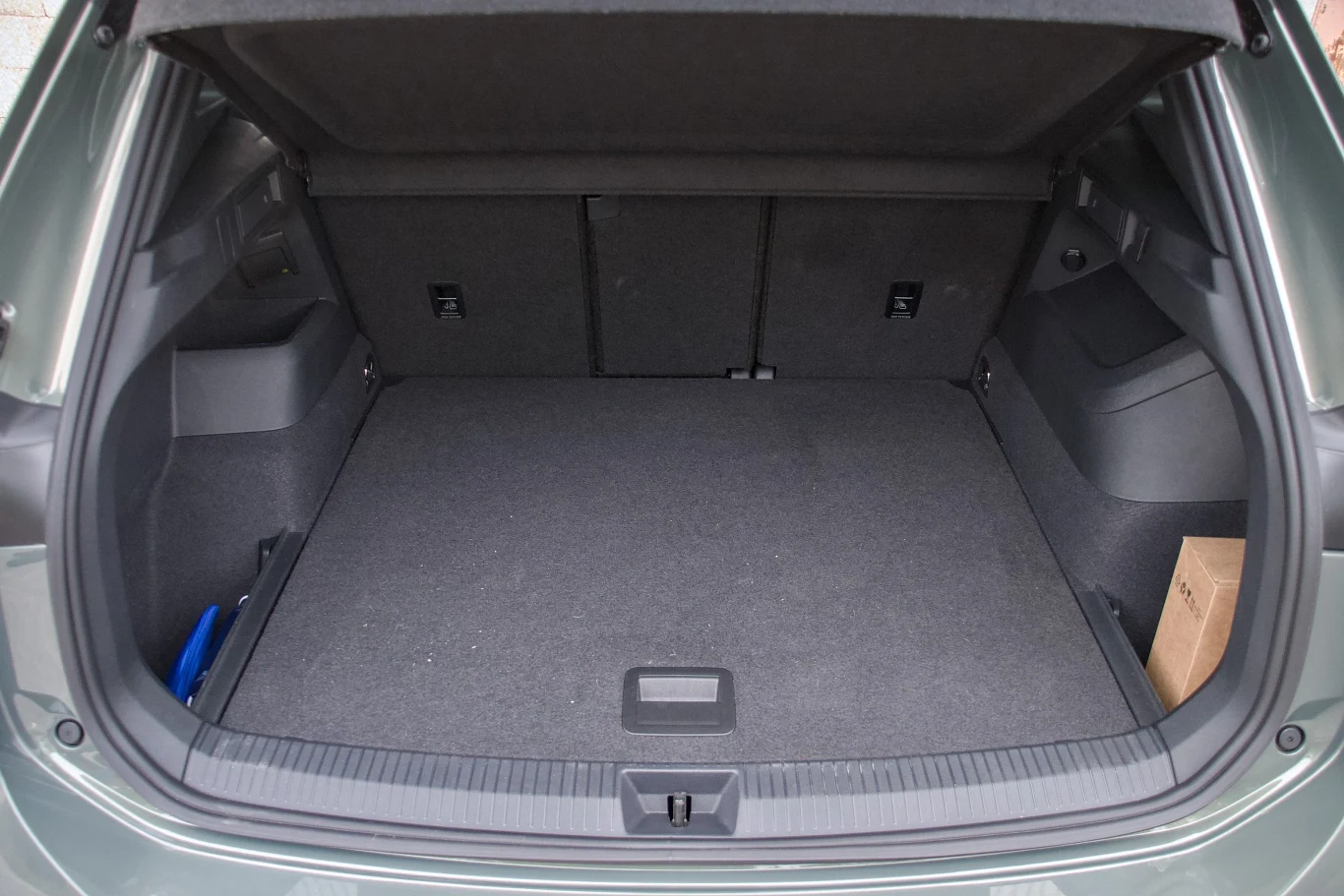 Pojemny bagażnik samochodu osobowego z rozłożoną tylną kanapą, wyłożony ciemną wykładziną, pusta przestrzeń gotowa do załadunku, po bokach widoczne są niewielkie wnęki i schowki, w rogu kartonowe opakowanie.