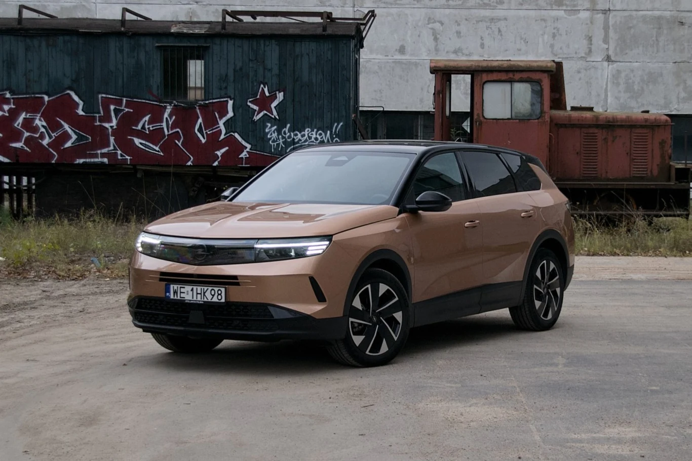 Brązowy samochód osobowy typu SUV stoi zaparkowany na utwardzonej powierzchni, w tle znajduje się stary wagon kolejowy i lokomotywa z widocznym graffiti na ścianie wagonu oraz budynek o betonowej strukturze.