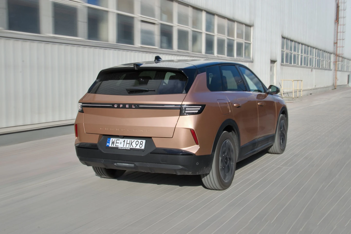 Brązowy samochód marki Opel, model SUV, porusza się po miejskiej ulicy obok długiego, przemysłowego budynku z oknami.