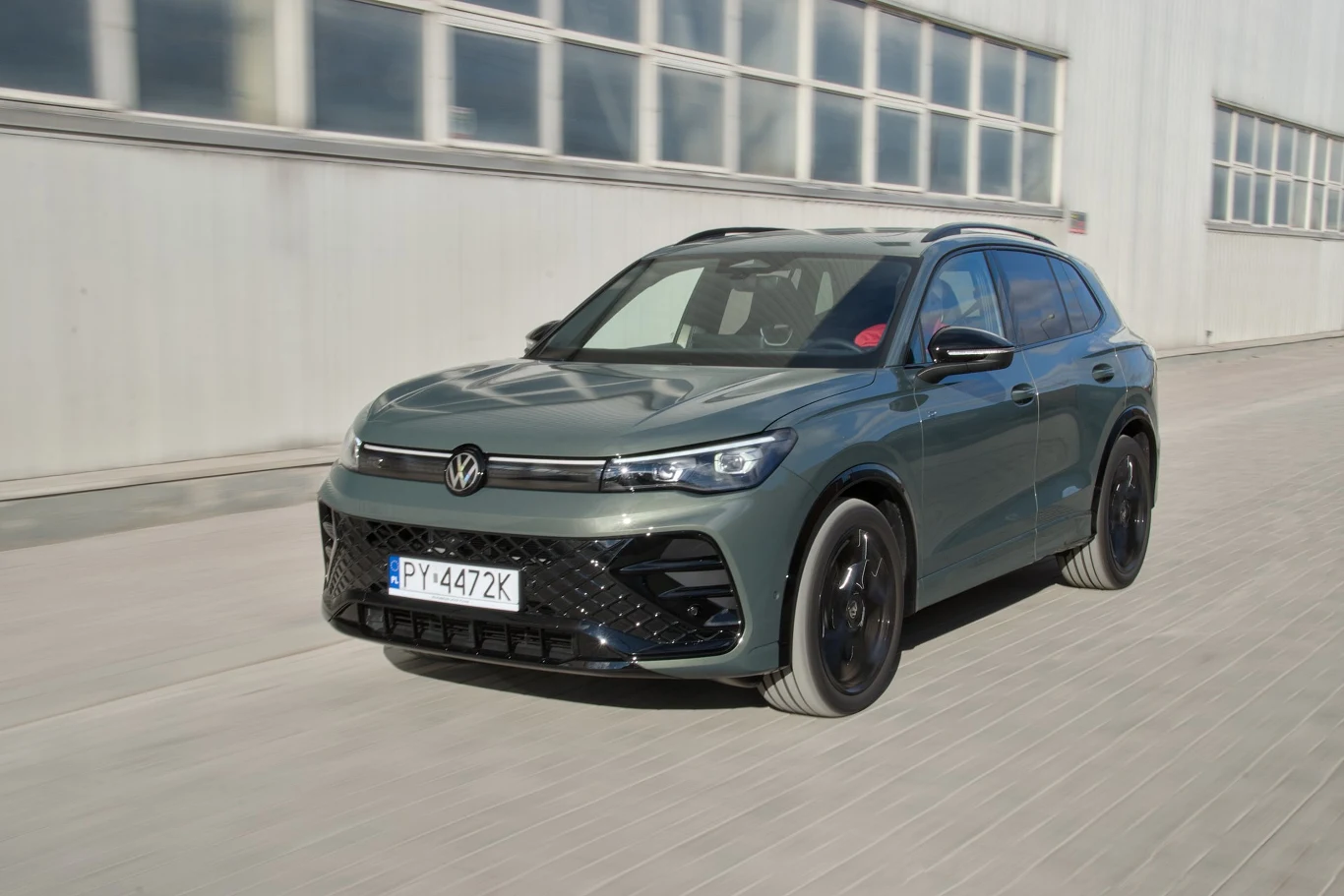 Nowoczesny SUV marki Volkswagen poruszający się po betonowej nawierzchni przy fasadzie dużego przemysłowego budynku. Pojazd w zielonkawym kolorze z czarnymi felgami i tablicą rejestracyjną PY 4472K, dynamiczny wygląd i linia auta podkreślają jego sport...